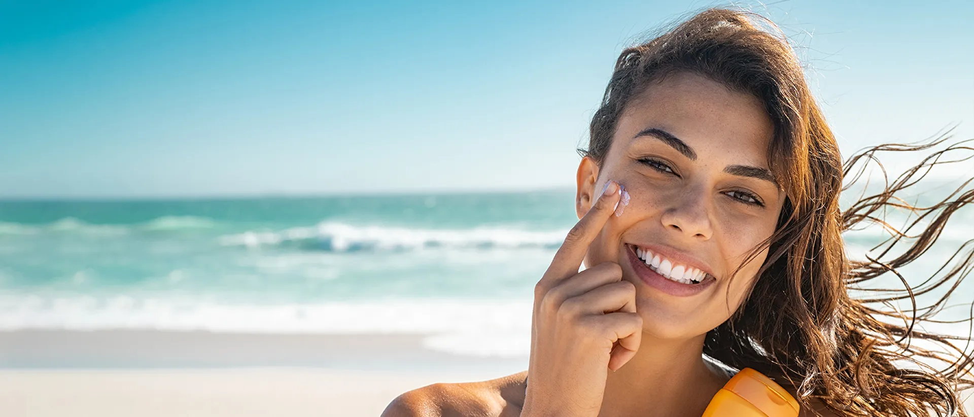 Woman on a beach applying sunscreen on her face - summer skin care routine for oily skin.