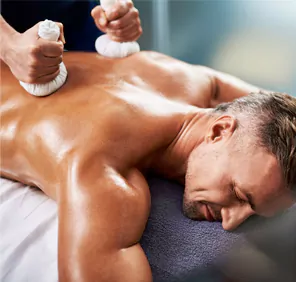 Two white cloth pouches are placed on a man's back during ayurvedic massage therapy.
