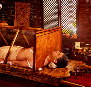 A man is sleeping on a wooden box table for Swedana Ayurveda therapy.
