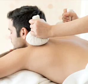 A person's hands hold two white cloth bundles against a man's bare back during an ayurvedic body massage.
